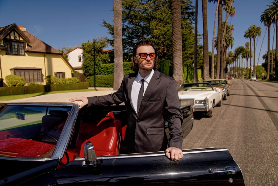 The black Cadillac DeVille with its door open on a street lined with palms and classic Cadillacs