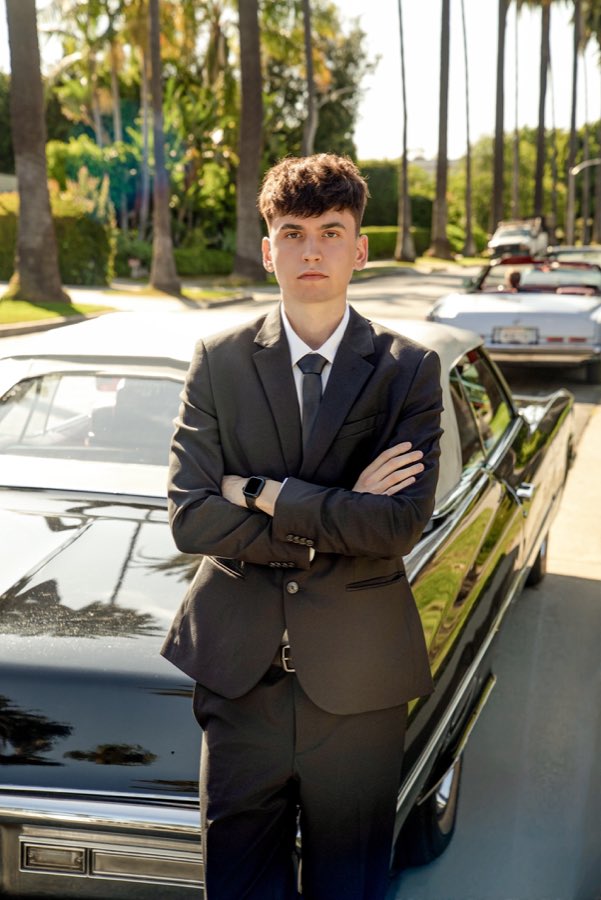 Young man in a suit standing by the black Cadillac, a white convertible parked behind