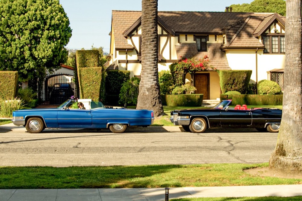 The blue and the black 1970 Cadillac DeVille convertibles, roof down, parked on a palm-lined Los Angeles street