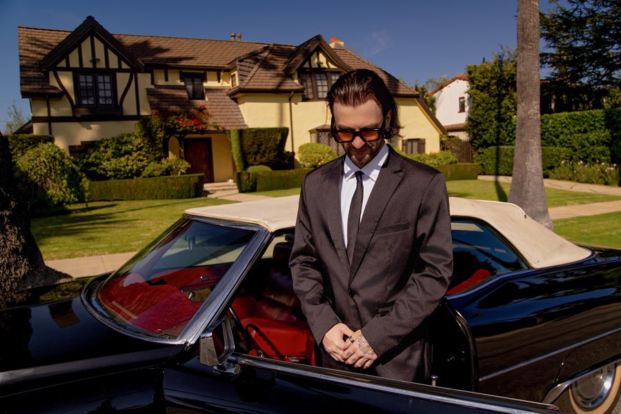 The black 1970 Cadillac DeVille parked outside a Tudor-style house in Los Angeles, a man in a dark suit beside it
