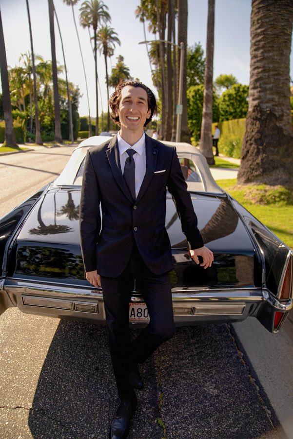 Young man in a dark suit at the back of the black Cadillac DeVille on a palm-lined street