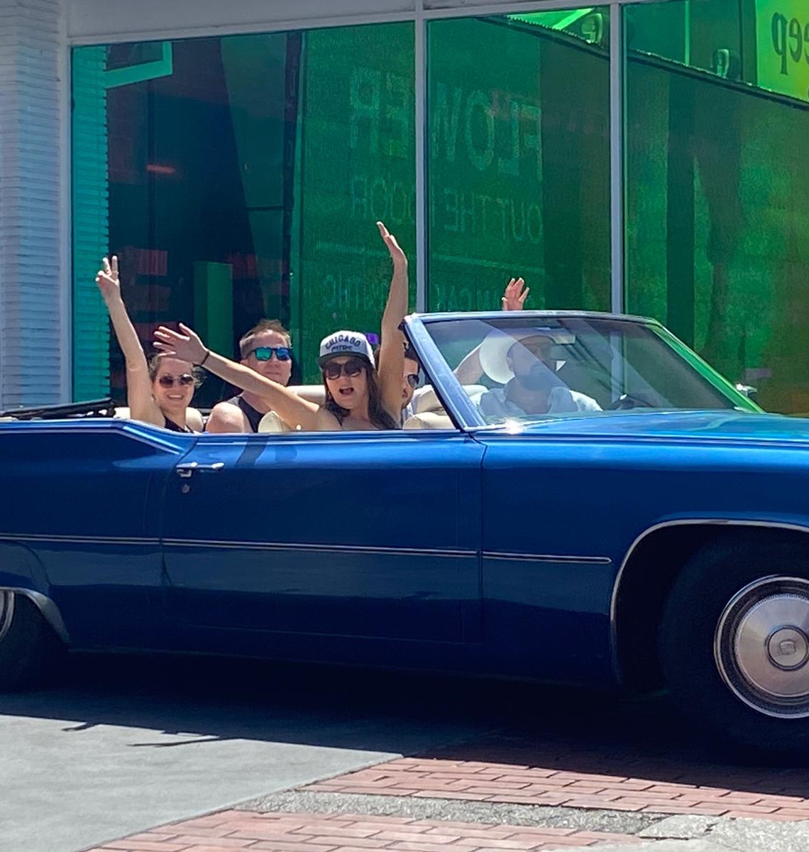 People waving from the blue 1970 Cadillac DeVille convertible, roof down, on a sunny Los Angeles street