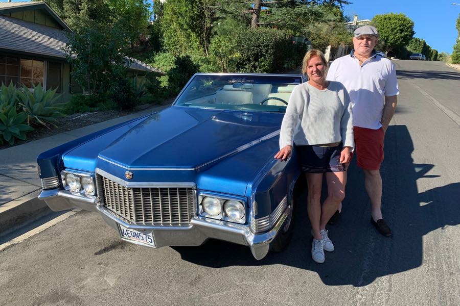 Blue 1970 Cadillac DeVille convertible parked on a sunny residential street, two people standing beside it