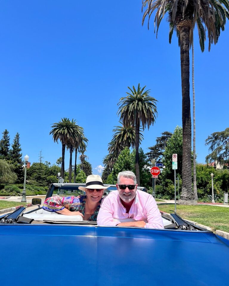 Two guests smiling from the blue Cadillac convertible on a palm-lined Beverly Hills street