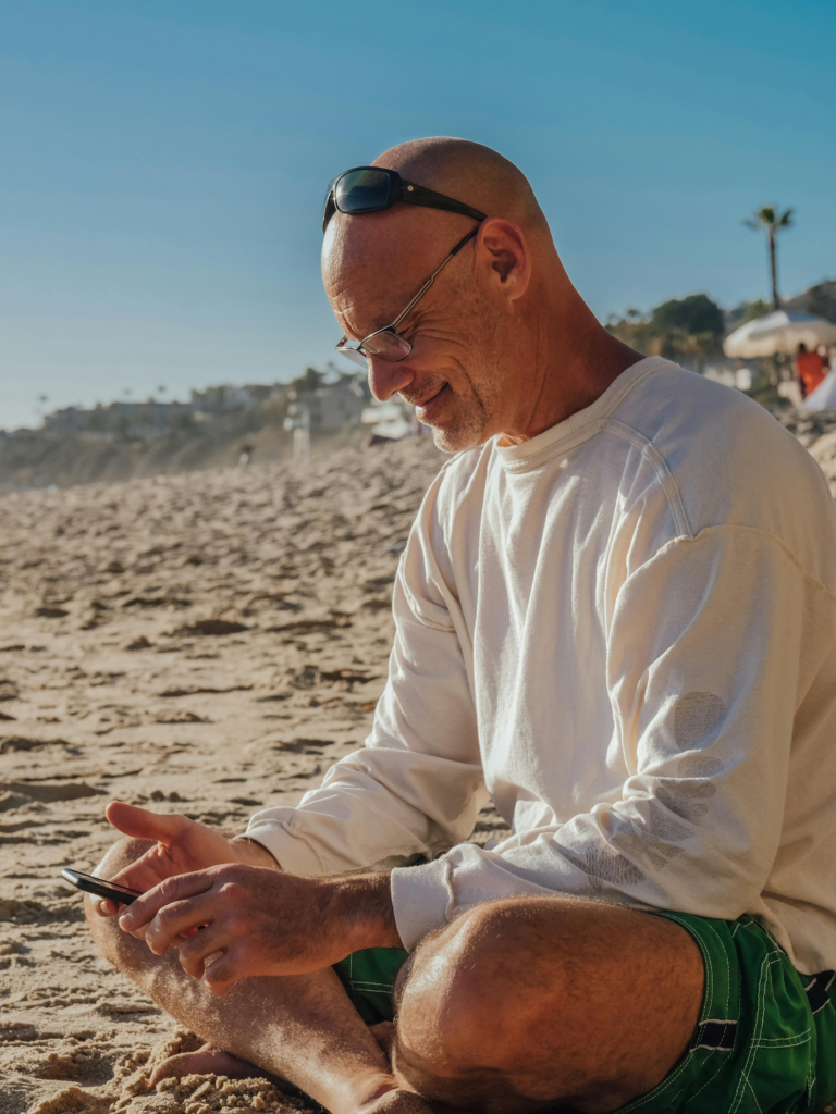 A man sitting cross-legged on the sand looking at his phone