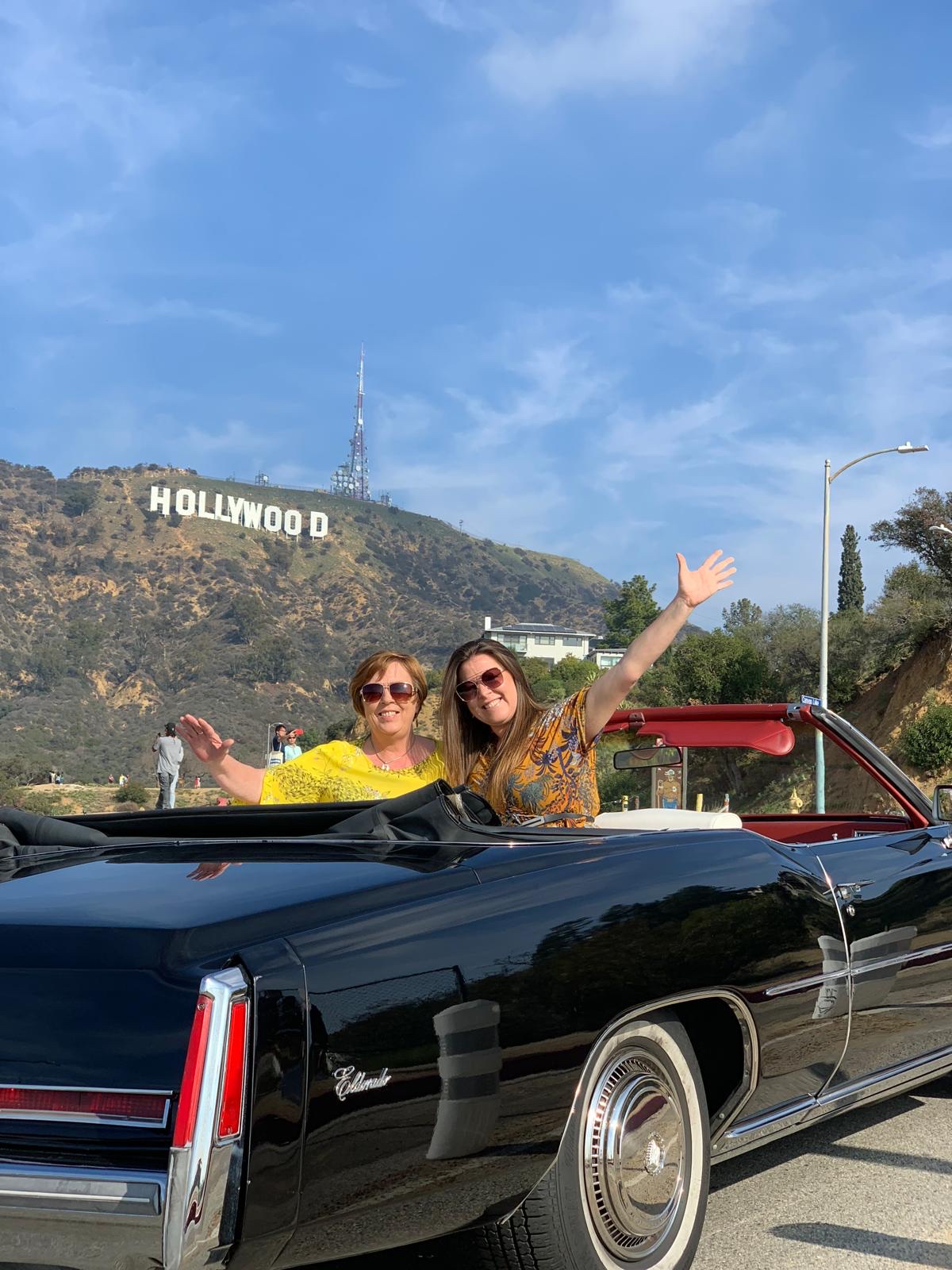 Two Guests Waving From A Black Classic Cadillac Convertible Below The Hollywood Sign